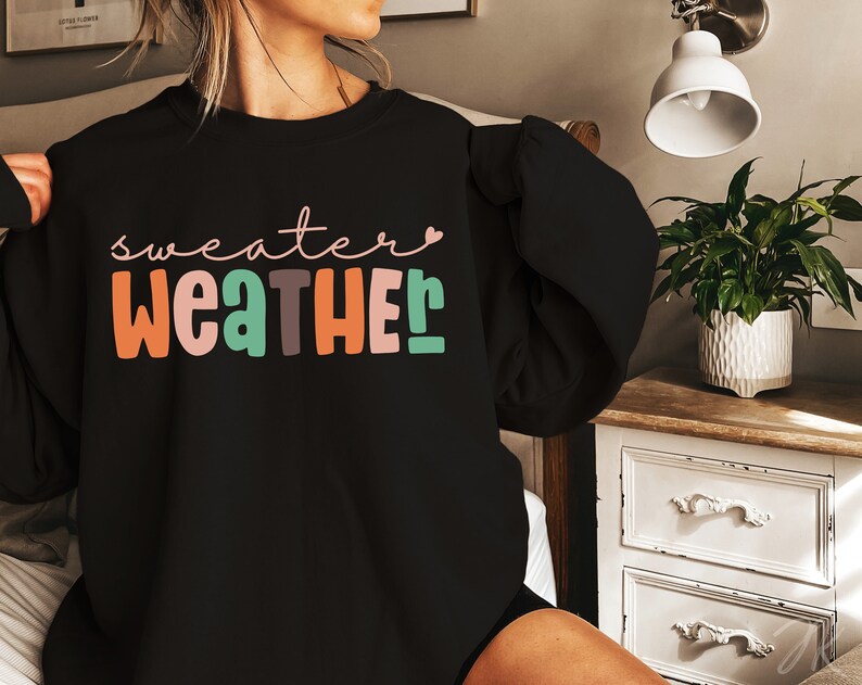 以下が含まれることがあります： カラフルで遊び心のある文字で「sweater weather」と書かれた黒いスウェットシャツ。テキストにはオレンジ、ピンク、グリーン、ブラウンの色が含まれています。「sweater」という単語は筆記体で書かれており、「r」の上には小さなピンク色のハートがあります。