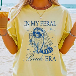 May include: Yellow t-shirt with the text "IN MY FERAL Bride ERA" in navy blue. The graphic features a raccoon wearing heart-shaped sunglasses, a veil, and holding a champagne glass. The raccoon is surrounded by stars.