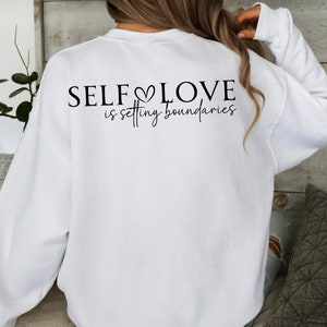 Puede incluir: Sudadera blanca con el texto "SELF LOVE is setting boundaries" impreso en la espalda.