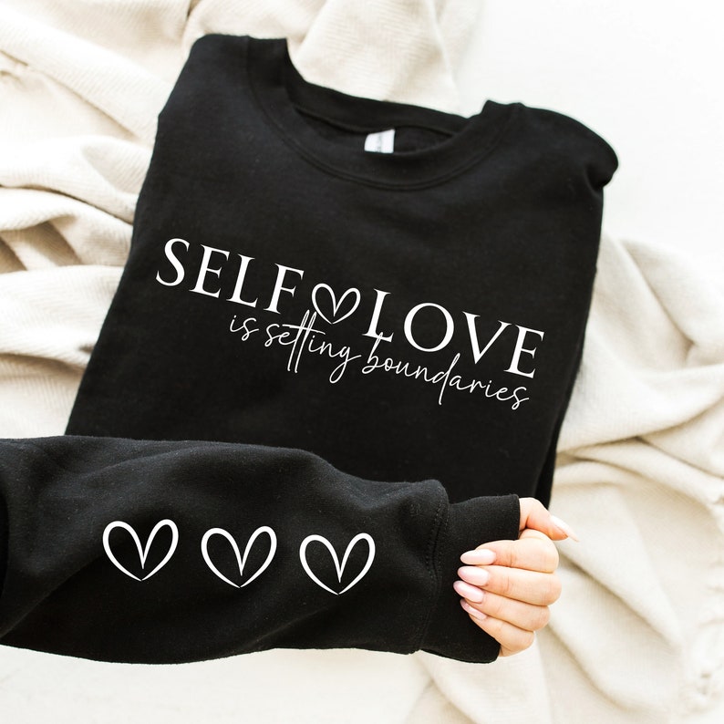Puede incluir: Sudadera negra con el texto "SELF LOVE is setting boundaries" en letras blancas con un s&iacute;mbolo de coraz&oacute;n. La sudadera tiene tres s&iacute;mbolos de coraz&oacute;n blancos en la manga.