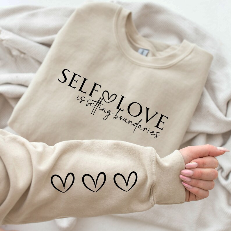Puede incluir: Una sudadera beige con el texto "SELF LOVE is setting boundaries" y tres peque&ntilde;os corazones en la manga.