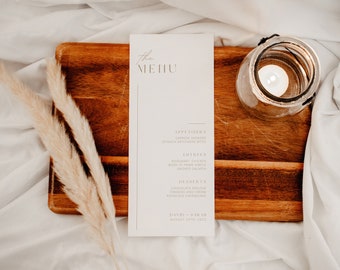Puede incluir: Una tarjeta de menú blanca con un borde dorado y la palabra "the MENU" en la parte superior. El menú enumera aperitivos, platos principales y postres. La tarjeta está sobre una tabla de cortar de madera con una vela en un frasco de vidrio.