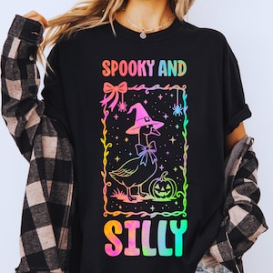 May include: Black t-shirt with a colorful graphic that reads "Spooky and Silly." The design features a goose wearing a witch hat and bow tie, a jack-o'-lantern, and spiders, perfect for Halloween.