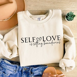 Puede incluir: Una sudadera de color crema con el texto "SELF LOVE is setting boundaries" en letras negras. La sudadera est&aacute; colocada sobre una superficie de madera con un sombrero marr&oacute;n, unos vaqueros azules y una peque&ntilde;a planta en maceta.
