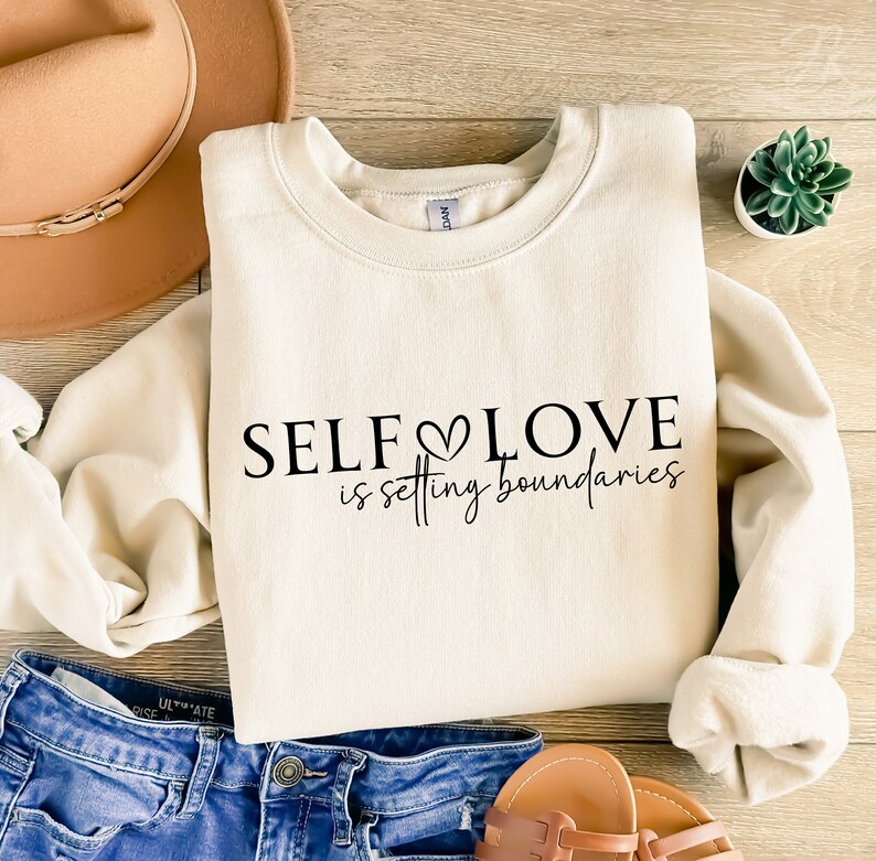 Puede incluir: Una sudadera de color crema con el texto "SELF LOVE is setting boundaries" en letras negras. La sudadera est&aacute; colocada sobre una superficie de madera con un sombrero marr&oacute;n, unos vaqueros azules y una peque&ntilde;a planta en maceta.