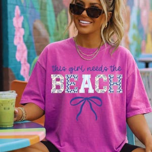 Op de afbeelding: Een magenta T-shirt met de tekst "this girl needs the BEACH" in blauw en wit. Het woord "BEACH" heeft een blauw-wit schelpenpatroon. Een blauwe strik-afbeelding staat eronder. Het shirt wordt door een persoon gedragen.