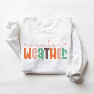 以下が含まれることがあります： レトロなフォントで「sweater weather」と書かれた白いスウェットシャツ。テキストはオレンジ、ピンク、緑、茶色です。秋にぴったりの長袖トップスです。