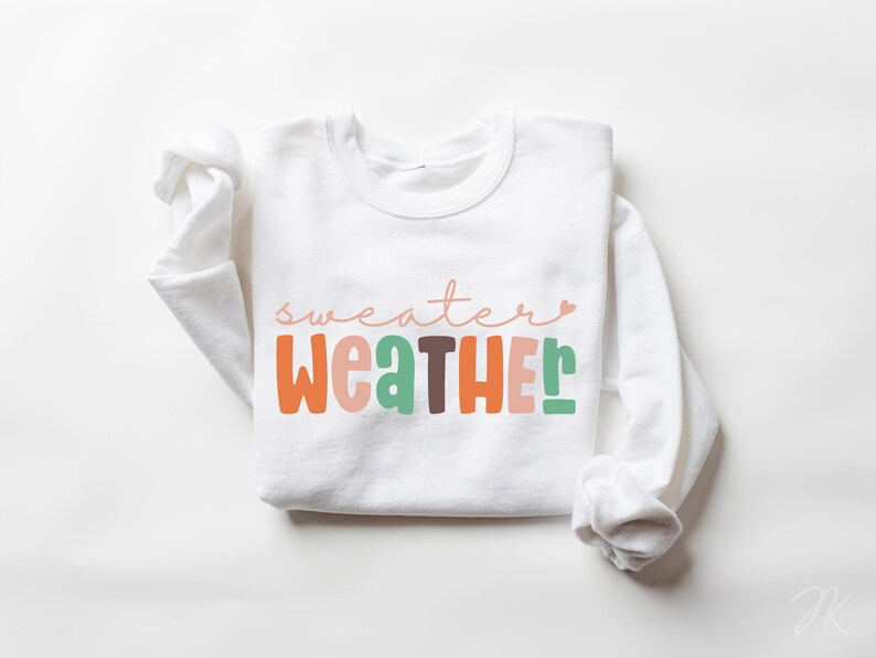 以下が含まれることがあります： レトロなフォントで「sweater weather」と書かれた白いスウェットシャツ。テキストはオレンジ、ピンク、緑、茶色です。秋にぴったりの長袖トップスです。
