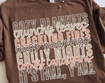 May include: A brown t-shirt with the text "COZY BLANKETS, CRUNCHY LEAVES, CRACKLING FIRES, CRISP MORNINGS, CHILLY NIGHTS, COMFY CARDIGANS, IT'S FALL, Y'ALL" in a layered design. The shirt has a crew neck and short sleeves.