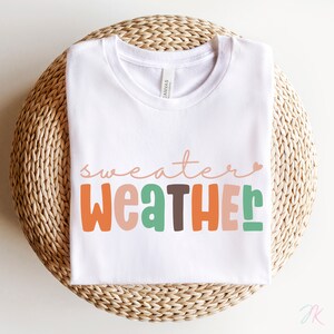 以下が含まれることがあります： カラフルで遊び心のある文字で「sweater weather」と書かれた白いTシャツ。オレンジ、緑、茶色、ピンクの色合いが含まれています。シャツは、織られた円形の表面に折りたたまれています。