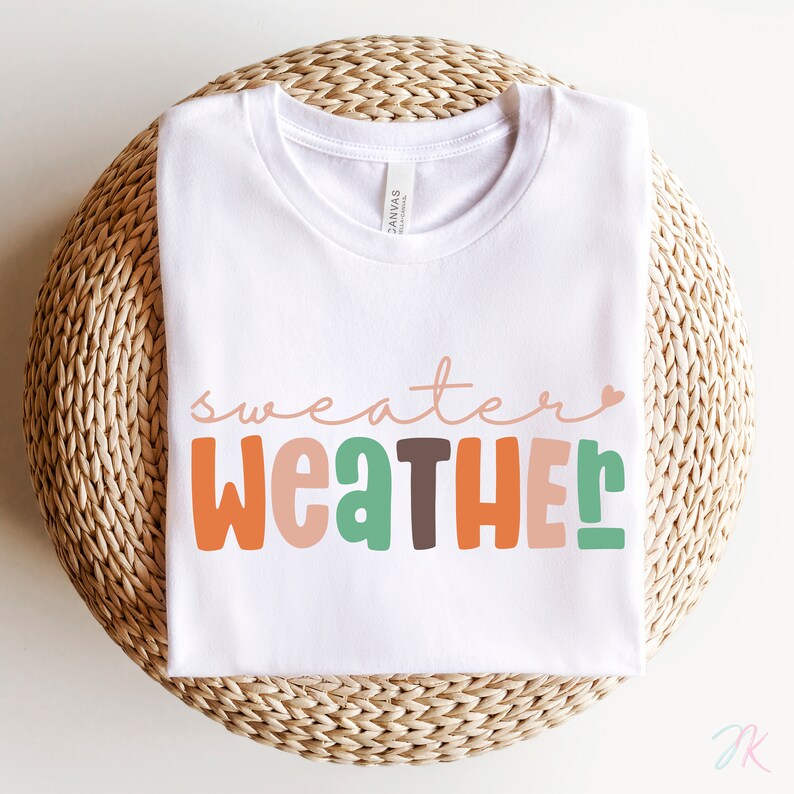 以下が含まれることがあります： カラフルで遊び心のある文字で「sweater weather」と書かれた白いTシャツ。オレンジ、緑、茶色、ピンクの色合いが含まれています。シャツは、織られた円形の表面に折りたたまれています。