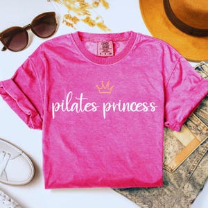 Puede incluir: Una camiseta rosa intenso con la frase "pilates princess" en escritura blanca y una corona dorada. La camiseta tiene cuello redondo y mangas enrolladas. Gafas de sol y un sombrero en el fondo.