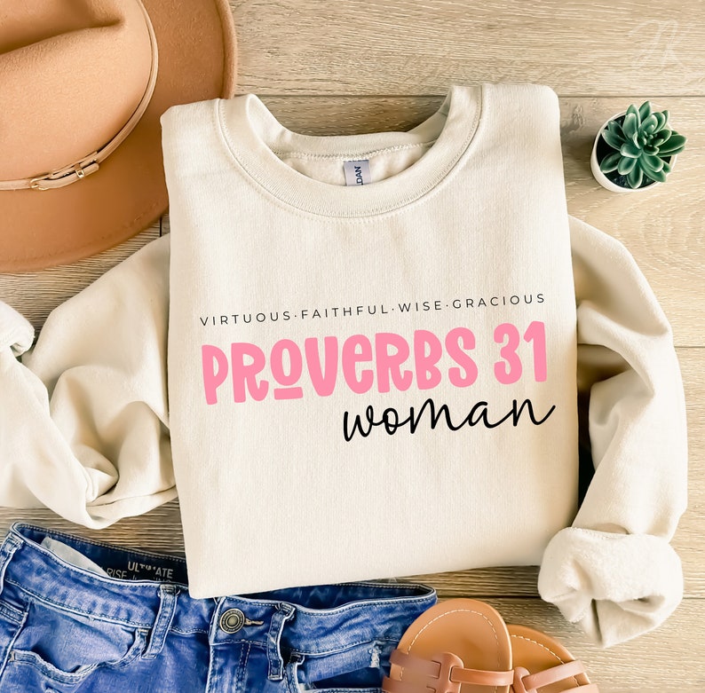 K&ouml;nnte beinhalten: Ein beiger Pullover mit dem Aufdruck "VIRTUOUS-FAITHFUL-WISE-GRACIOUS PROVERBS 31 woman" in Pink und Schwarz.