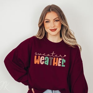 以下が含まれることがあります： カラフルで遊び心のある文字で「Sweater Weather」と書かれた、バーガンディ色のスウェットシャツ。テキストは気まぐれなフォントで、各文字は異なる色です。スウェットシャツは無地で、クルーネックです。