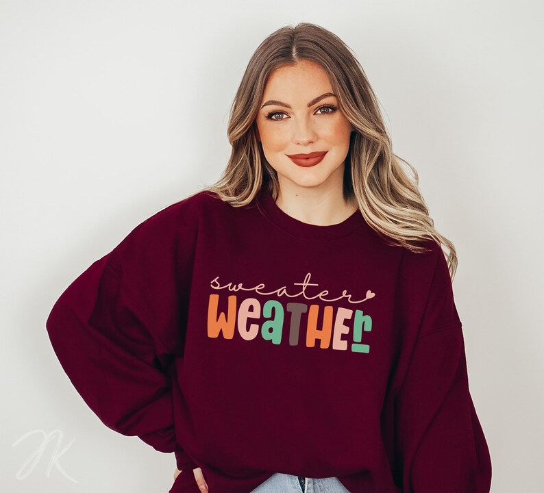 以下が含まれることがあります： カラフルで遊び心のある文字で「Sweater Weather」と書かれた、バーガンディ色のスウェットシャツ。テキストは気まぐれなフォントで、各文字は異なる色です。スウェットシャツは無地で、クルーネックです。