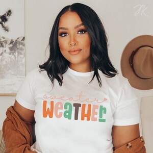 以下が含まれることがあります： カラフルで遊び心のある文字で「sweater weather」と書かれた白いTシャツ。 シャツはクルーネックと半袖です。 茶色のジャケットが肩にかけられています。 背景には茶色の帽子が掛かっています。
