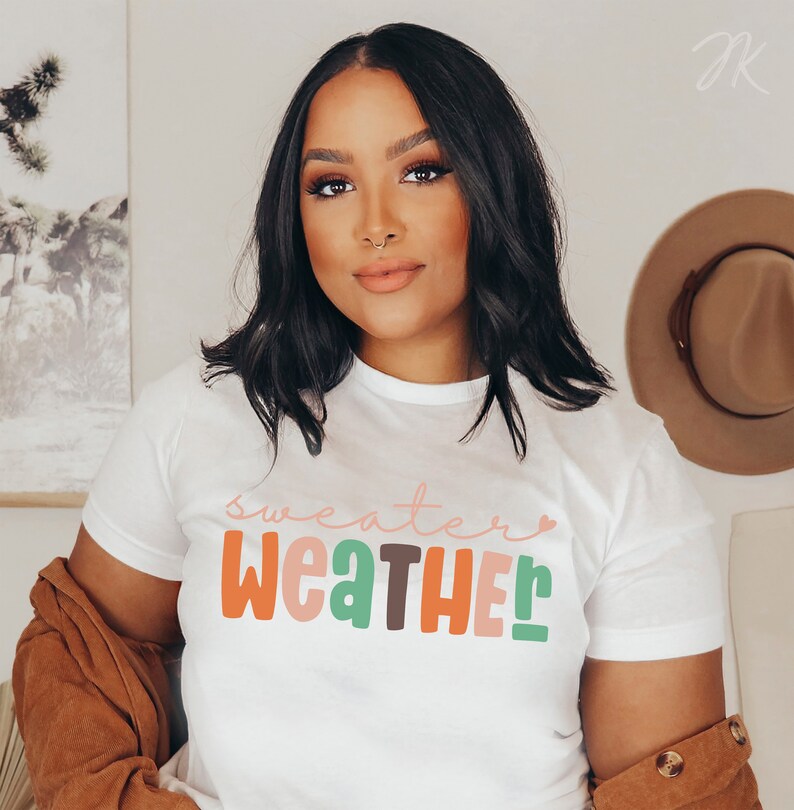 以下が含まれることがあります： カラフルで遊び心のある文字で「sweater weather」と書かれた白いTシャツ。 シャツはクルーネックと半袖です。 茶色のジャケットが肩にかけられています。 背景には茶色の帽子が掛かっています。