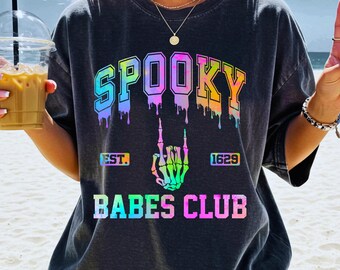 May include: Dark gray t-shirt with the word "SPOOKY" in a rainbow dripping font. Below the word is a skeleton hand making a rock on gesture. The text "EST. 1629" and "BABES CLUB" are also on the shirt.