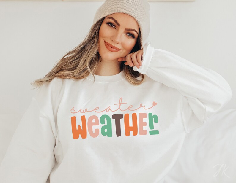 以下が含まれることがあります： カラフルで遊び心のある文字で「Sweater Weather」と書かれた白いスウェットシャツ。テキストには「Weather」という単語の上にピンクのハートが含まれています。スウェットシャツは長袖で、クルーネックです。