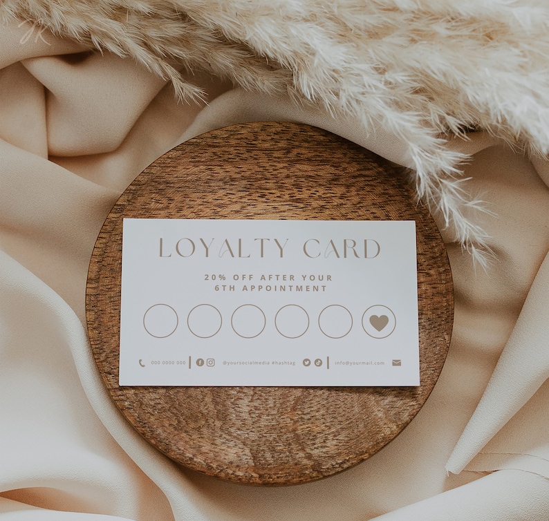 Op de afbeelding: Een witte klantenkaart met de tekst "LOYALTY CARD" en "20% OFF AFTER YOUR 6TH APPOINTMENT" gedrukt in een lichtbruine kleur. De kaart heeft zes cirkels en een hartsymbool. De kaart ligt op een houten bord.