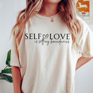 Puede incluir: Una camiseta beige con el texto "SELF LOVE is setting boundaries" en letras negras. El texto incluye un peque&ntilde;o s&iacute;mbolo de coraz&oacute;n.