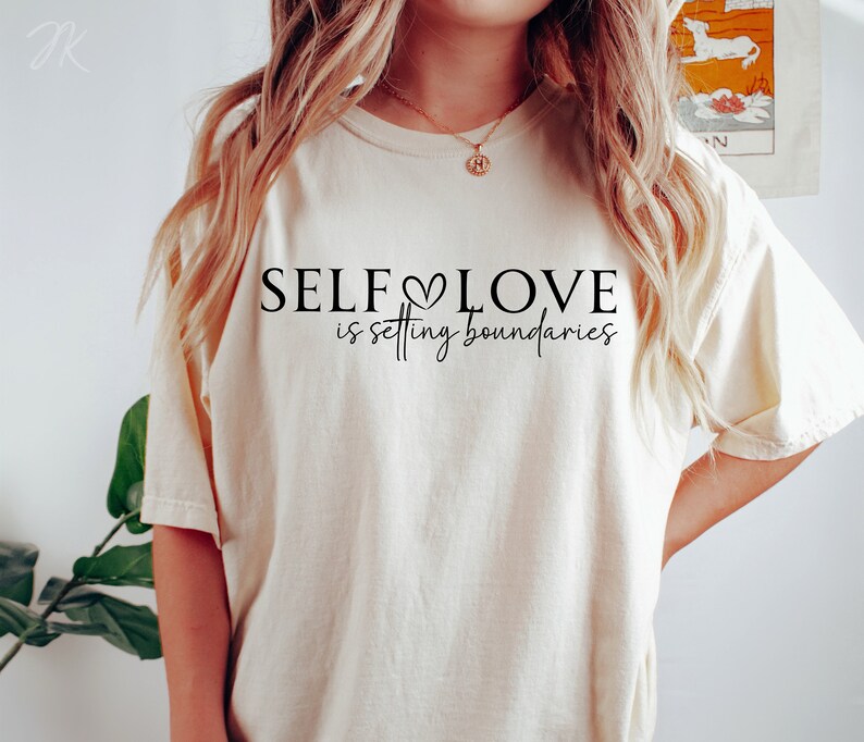 Puede incluir: Una camiseta beige con el texto "SELF LOVE is setting boundaries" en letras negras. El texto incluye un peque&ntilde;o s&iacute;mbolo de coraz&oacute;n.