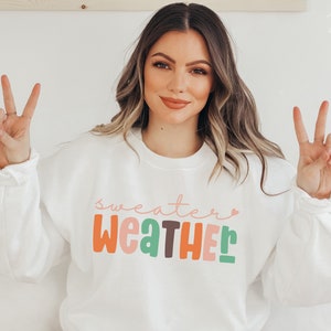 以下が含まれることがあります： カラフルで遊び心のある文字で「Sweater Weather」と書かれた白いスウェットシャツ。スウェットシャツはクルーネックと長袖です。モデルは両手でピースサインをしています。
