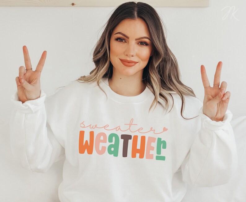 以下が含まれることがあります： カラフルで遊び心のある文字で「Sweater Weather」と書かれた白いスウェットシャツ。スウェットシャツはクルーネックと長袖です。モデルは両手でピースサインをしています。