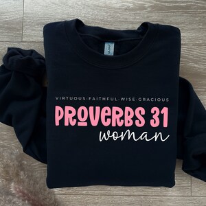 K&ouml;nnte beinhalten: Schwarzer Pullover mit dem Text "PROVERBS 31 woman" in rosa Schrift. Der Text "VIRTUOUS. FAITHFUL. WISE. GRACIOUS" ist in wei&szlig; &uuml;ber dem Haupttext gedruckt.