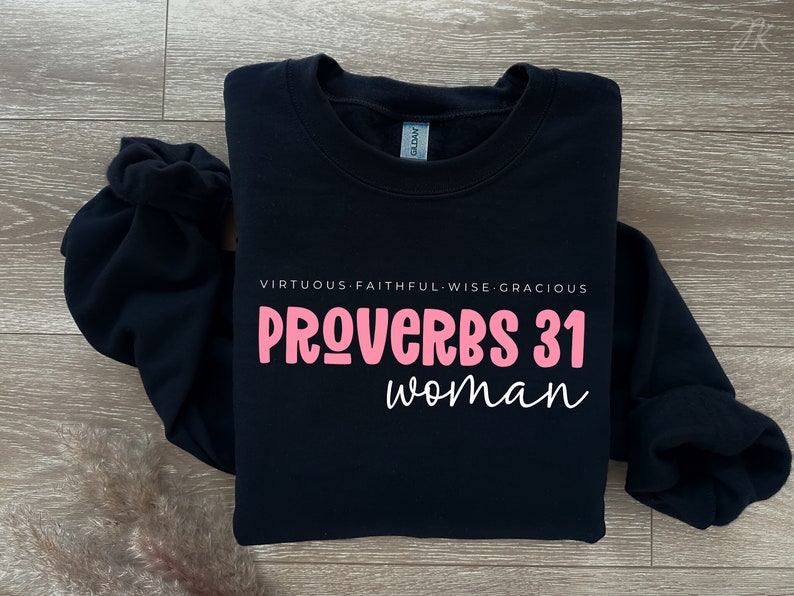 K&ouml;nnte beinhalten: Schwarzer Pullover mit dem Text "PROVERBS 31 woman" in rosa Schrift. Der Text "VIRTUOUS. FAITHFUL. WISE. GRACIOUS" ist in wei&szlig; &uuml;ber dem Haupttext gedruckt.