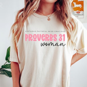 K&ouml;nnte beinhalten: Ein wei&szlig;es T-Shirt mit dem Text "SPR&Uuml;CHE 31 Frau" in rosa und schwarzer Schrift. Der Text "TUGENDHAFT - TREU - WEISE - GN&Auml;DIG" steht in Schwarz &uuml;ber dem Haupttext.