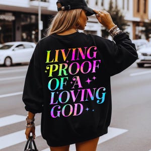 May include: Black sweatshirt with the text "LIVING PROOF OF A LOVING GOD" in a vibrant, rainbow-colored font. The sweatshirt is a crew neck style and is being worn by a person.