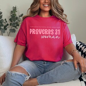 K&ouml;nnte beinhalten: Ein pinkfarbenes T-Shirt mit dem Text "VIRTUOUS. FAITHFUL. WISE. GRACIOUS. PROVERBS 31 woman" in pinker Schrift.