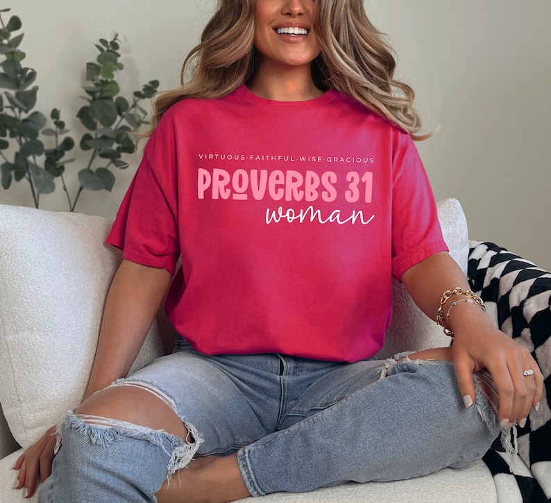 K&ouml;nnte beinhalten: Ein pinkfarbenes T-Shirt mit dem Text "VIRTUOUS. FAITHFUL. WISE. GRACIOUS. PROVERBS 31 woman" in pinker Schrift.
