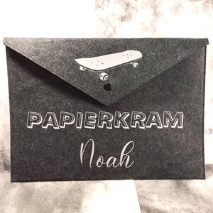 Könnte beinhalten: Dunkelgraue Filzumschlag mit weißer Skateboard-Grafik und dem Wort "PAPIERKRAM" in weißen, fetten Buchstaben. Der Name "Noah" ist darunter in Schreibschrift geschrieben. Der Umschlag hat einen Klappenverschluss mit einem schwarzen Druckknopf.