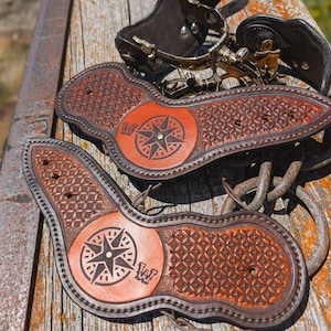 May include: A pair of brown leather spur straps featuring a stamped geometric pattern and a central circular medallion embossed with a compass rose design and the letters W. The straps include metal hardware and are constructed with dark stitching.