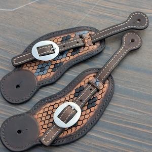 Puede incluir: Un par de correas de espuelas de cuero marrón con un patrón decorativo de escamas en tonos marrón, negro y azul. Cada correa tiene una hebilla plateada y costuras blancas. Diseñadas para uso ecuestre.