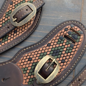 Puede incluir: Primer plano de dos correas de guitarra de cuero marrón con un patrón de escamas en tonos verdes, naranjas y negros. Cada correa presenta una hebilla de latón y costuras decorativas. Las correas están diseñadas para guitarras.