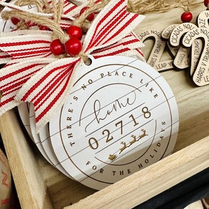 May include: A stack of white wooden Christmas ornaments with a rustic design. Each ornament has a circular shape with the text "There's no place like home for the holidays" and the number "02718". They are decorated with red and white striped ribbon and red beads.