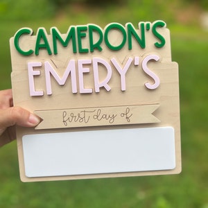 May include: Wooden sign with the text "Cameron's Emery's first day of" and a blank space for writing. The sign is decorated with green and pink letters.