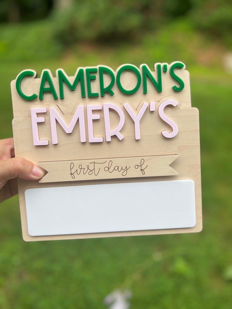 May include: Wooden sign with the text "Cameron's Emery's first day of" and a blank space for writing. The sign is decorated with green and pink letters.