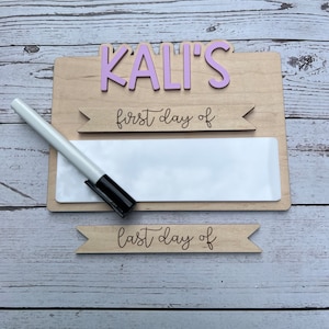 May include: A wooden sign with the name "KALI'S" in pink letters. The sign has a blank white space for writing with a dry erase marker. The sign also has two banners that say "first day of" and "last day of".