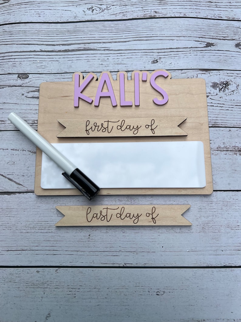 May include: A wooden sign with the name "KALI'S" in pink letters. The sign has a blank white space for writing with a dry erase marker. The sign also has two banners that say "first day of" and "last day of".