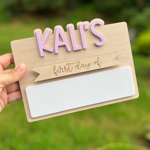 May include: A wooden sign with the name "KALI'S" in pink letters and a banner that says "first day of" in cursive. The sign has a blank white space for writing.