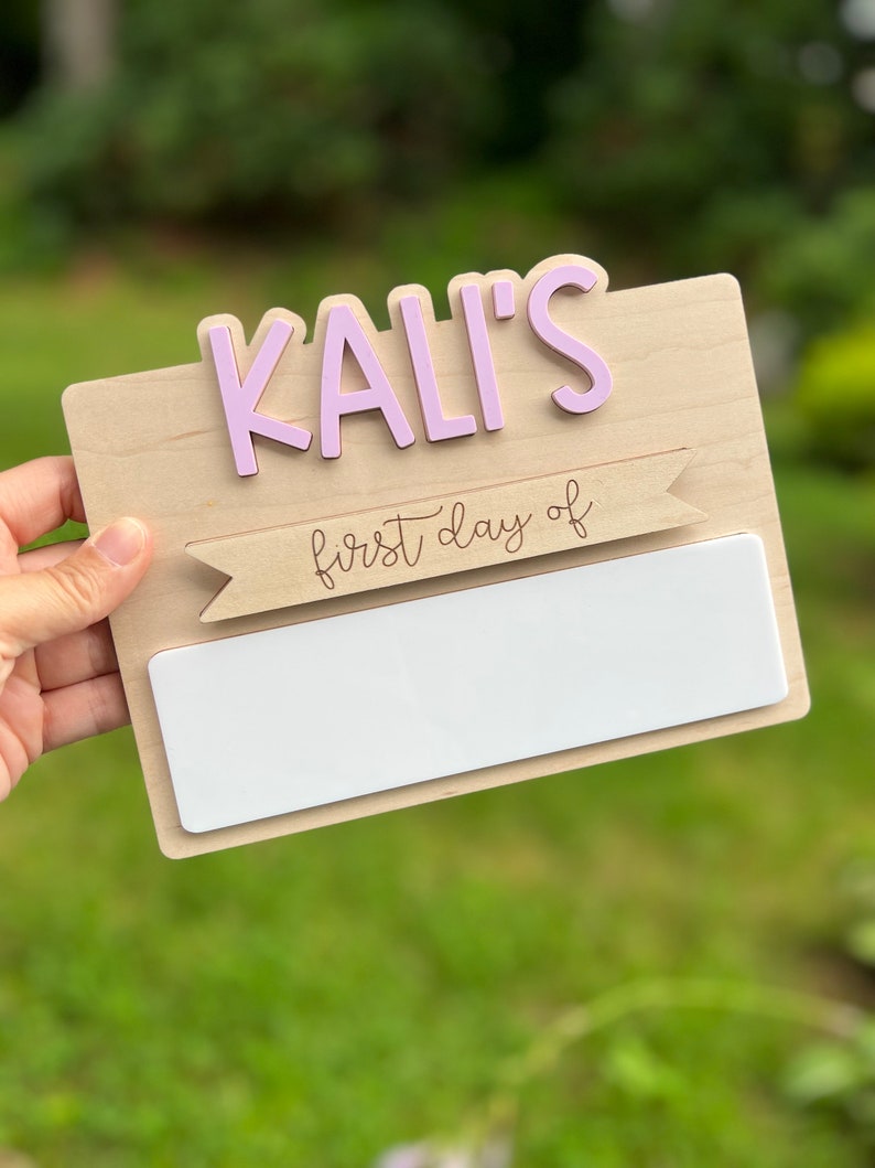 May include: A wooden sign with the name "KALI'S" in pink letters and a banner that says "first day of" in cursive. The sign has a blank white space for writing.