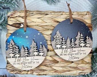 May include: Two round wooden ornaments with a blue and black starry night sky background. Each ornament features a silhouette of a forest with the text "I'll See You On The Other Side Of The Stars".
