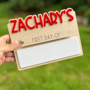 May include: A wooden sign with the name "Zachary's" in red letters. The sign also has a banner that says "First Day Of" and a blank space for writing.