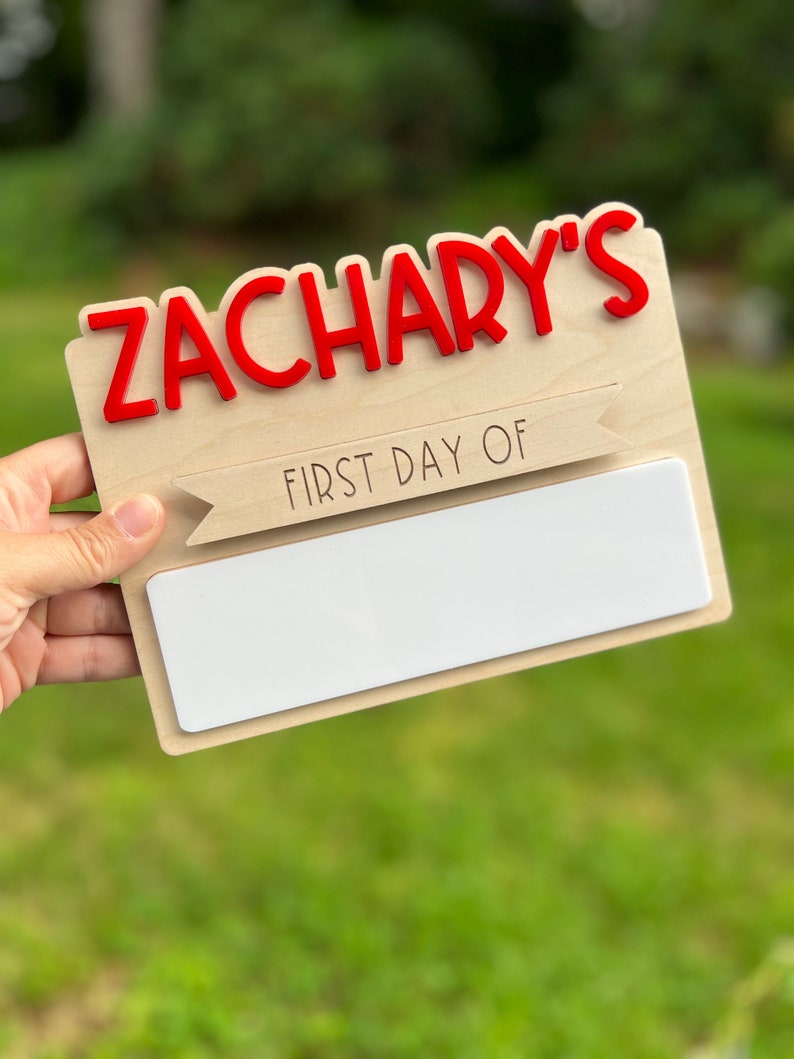 May include: A wooden sign with the name "Zachary's" in red letters. The sign also has a banner that says "First Day Of" and a blank space for writing.