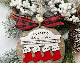 May include: A wooden Christmas ornament with a red and black buffalo plaid ribbon. The ornament is personalized with the text "The Leonards Merry Christmas 2022" and four stockings with the names "Eric", "Amy", "Carter", and "Charlie".