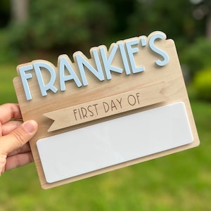May include: A wooden sign with the name "FRANKIE'S" in blue letters and a banner that says "FIRST DAY OF" with a blank space for writing. The sign is held in a hand.